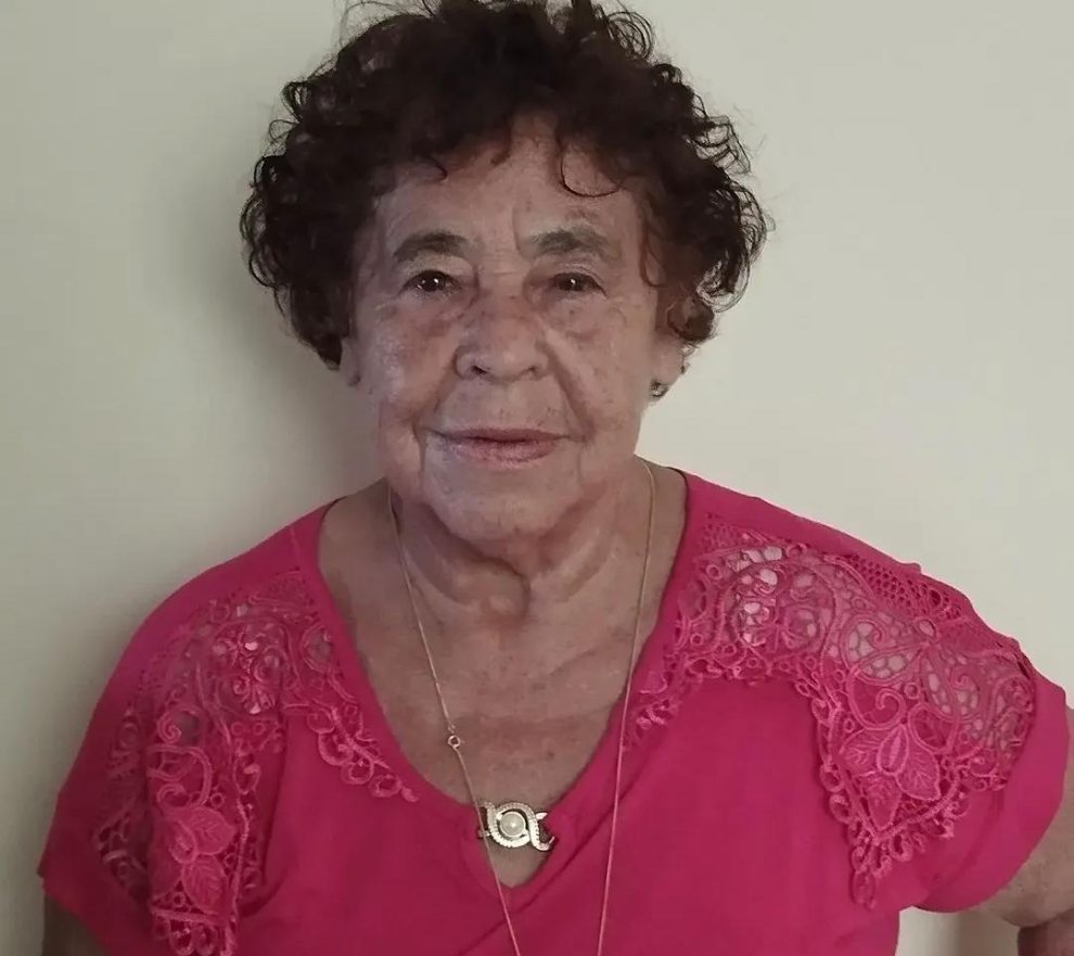 a foto mostra uma ideosa de 70 anos com blusa rosa, cabelos encaracolados e semblante levemente sorrindo